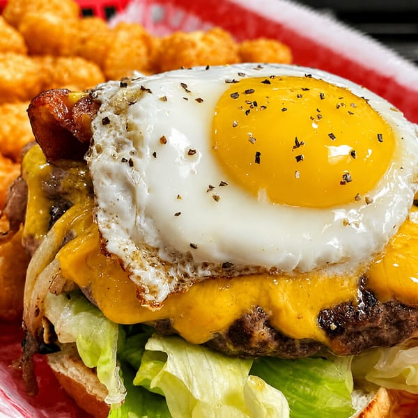 Close-up of a cheeseburger with bacon, fried egg, lettuce, and tater tots in a red basket.