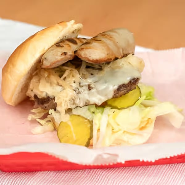 A Burgers & Brats burger with melted cheese, pickles, lettuce, and grilled bratwurst on a bun.
