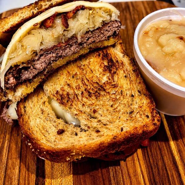 A grilled burger patty with melted cheese, sauerkraut, and bacon on toasted bread, with applesauce.