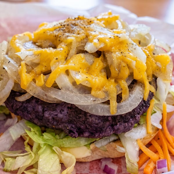 Close-up of a purple patty burger with melted cheese, grilled onions, lettuce, and shredded carrots.